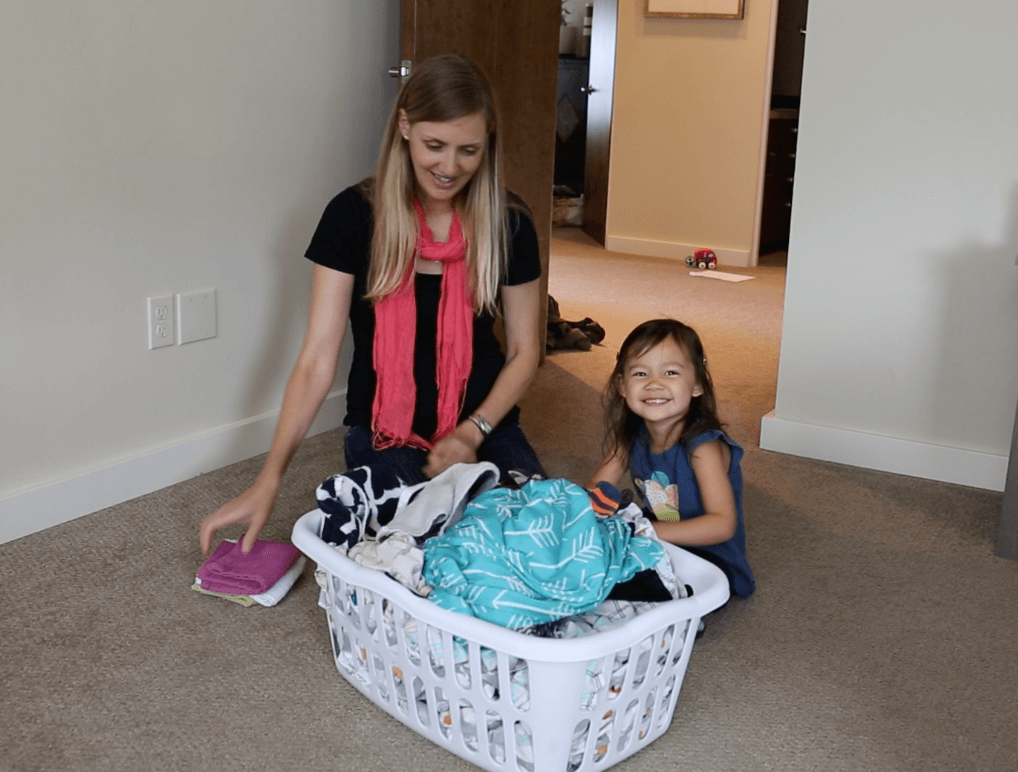 Child and adult with a full laundry basket, folding laundry. Child is smiling
