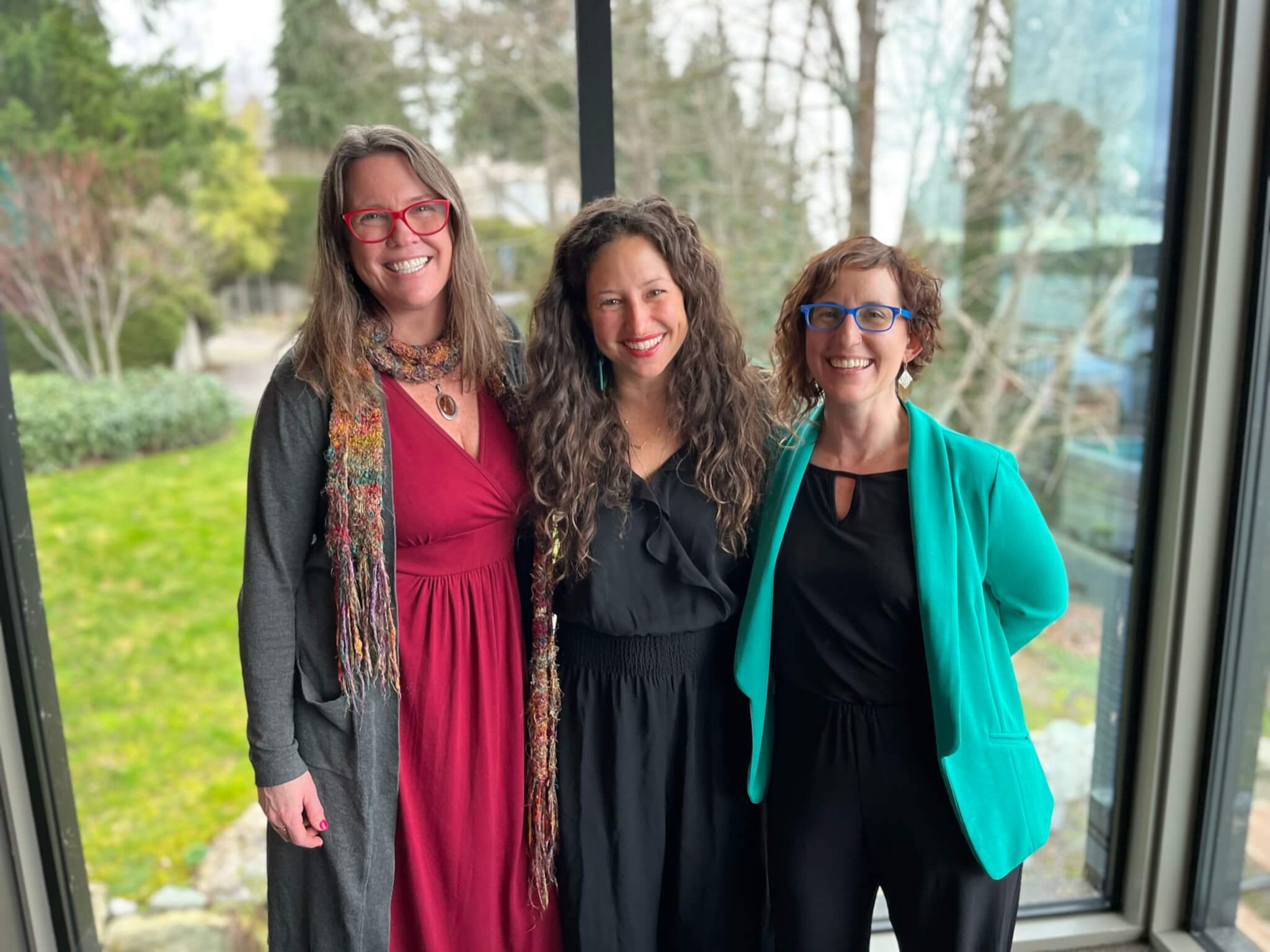 Three white women standing together in front of a window showing trees and grass.